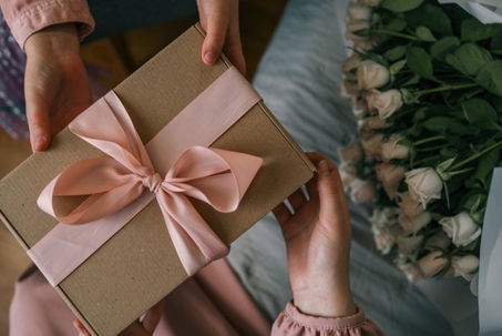 Hands of a child giving a wrapped gift with a satin ribbon to a woman holding a bouquet of roses, captured in warm indoor light. See less
